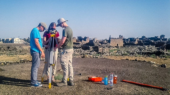 Three researchers use a surveying tripod on a hill in front of numerous ancient ruins.
